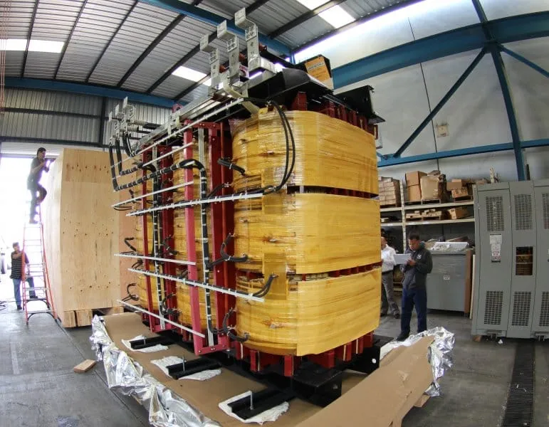 Large yellow and red electrical transformer wrapped in plastic in industrial warehouse with workers