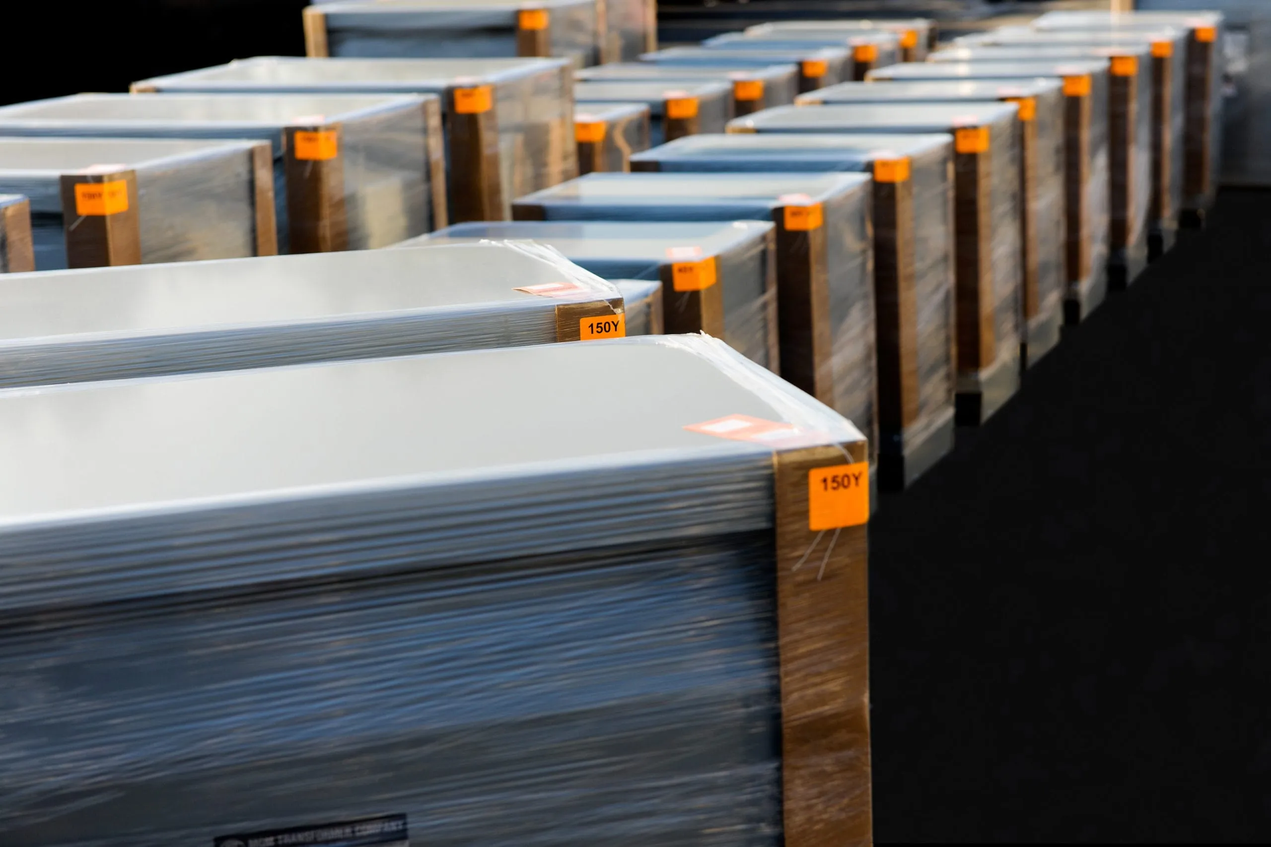Rows of wrapped aluminum transformer coils with orange inventory labels stacked in industrial warehouse