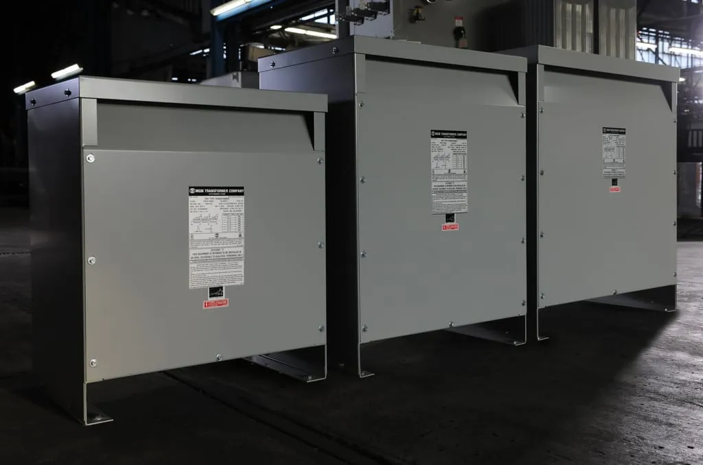Three industrial transformer units with gray enclosures and specification labels displayed in manufacturing facility