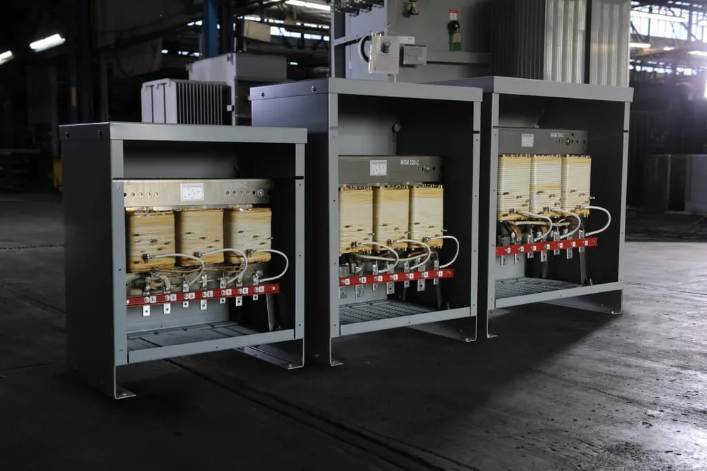 Three industrial power transformers with beige coils and red switching components in metal enclosures on factory floor