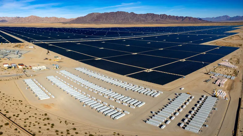 Large-scale solar farm with black photovoltaic panels and battery storage units in desert landscape with mountains