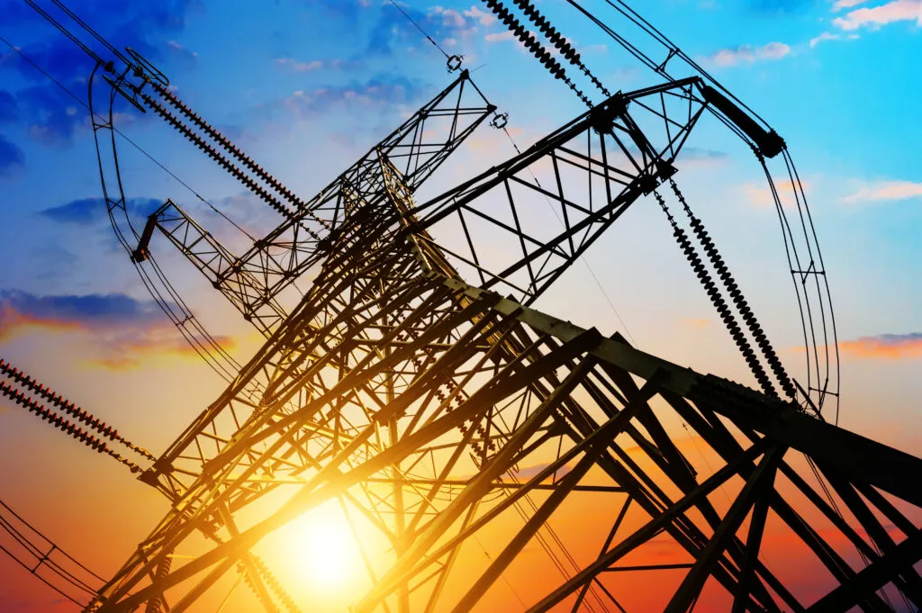 High-voltage transmission tower silhouetted against sunset sky with power lines, representing utility infrastructure and