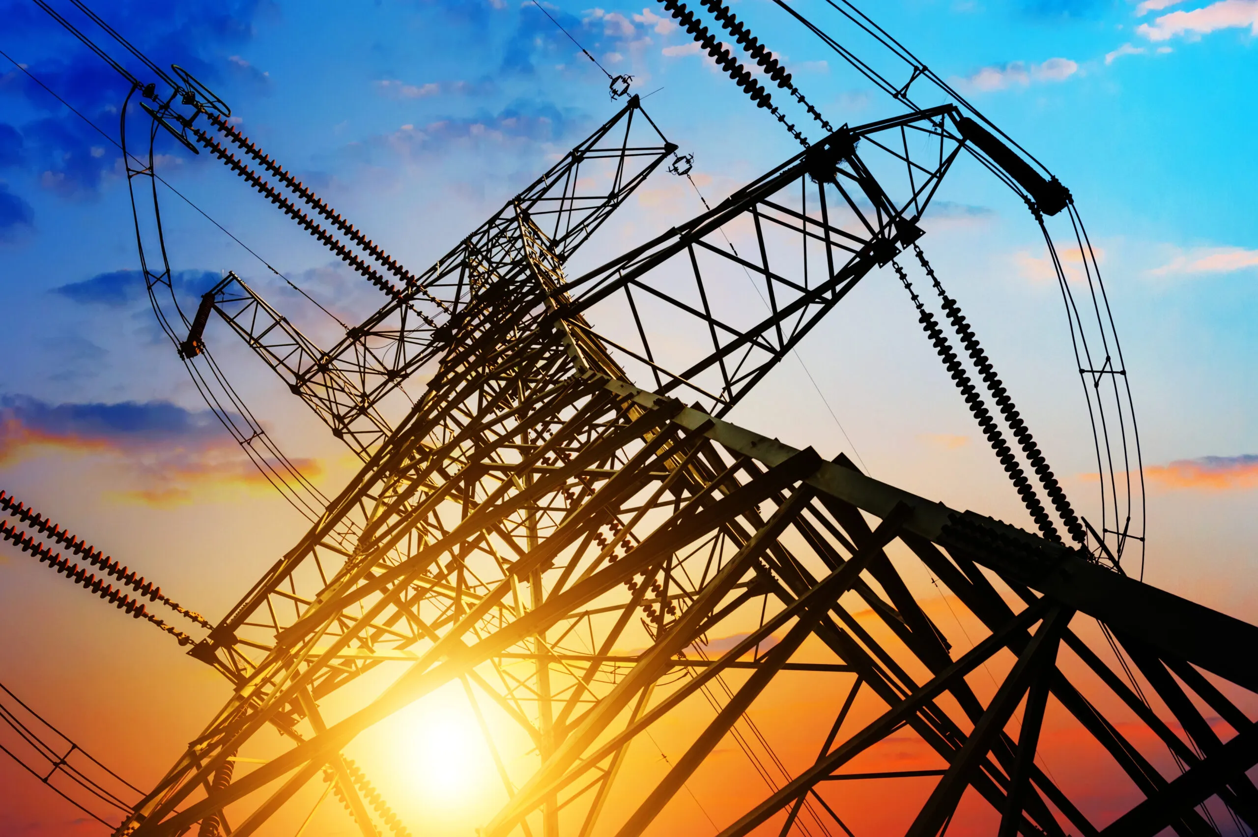 High-voltage transmission tower silhouetted against sunset sky with power lines, representing utility infrastructure and