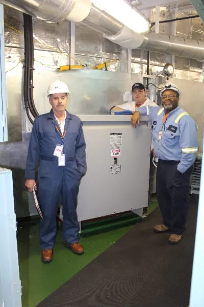 Three technicians in hard hats and safety gear pose beside industrial transformer equipment in factory setting